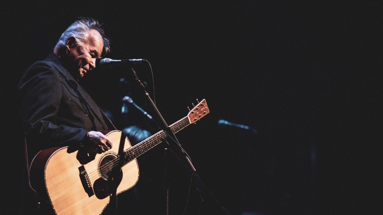 John Prine with a guitar in front of a microphone